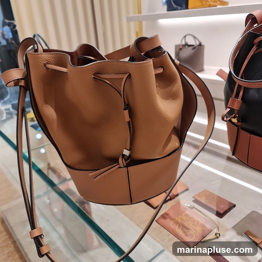 A close-up view of a tan leather bucket bag with drawstring closure, displayed on a glass shelf in a retail store. In the background, other handbags and small leather goods are visible, including a black and tan bag and a smaller grey handbag. The overall impression is of a luxury goods store display.