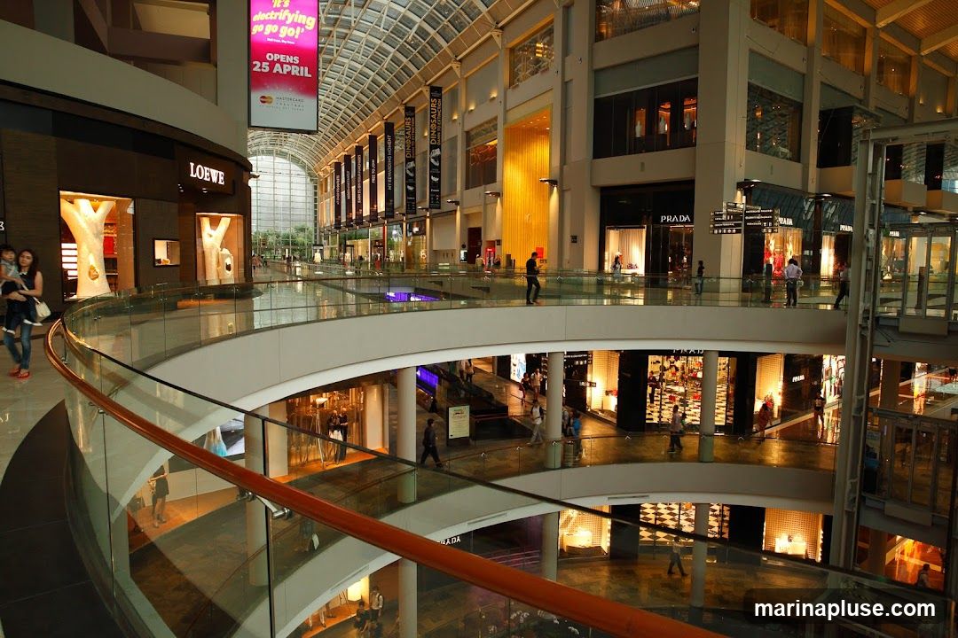 The image shows the interior of a large, modern shopping mall with multiple levels. There are various luxury brand stores visible, including Loewe and Prada. People are seen walking on the elevated walkways and browsing in the shops. The mall features a glass ceiling and a curved interior design with escalators and elevators. A large banner advertising an event opening on April 25th is displayed prominently.