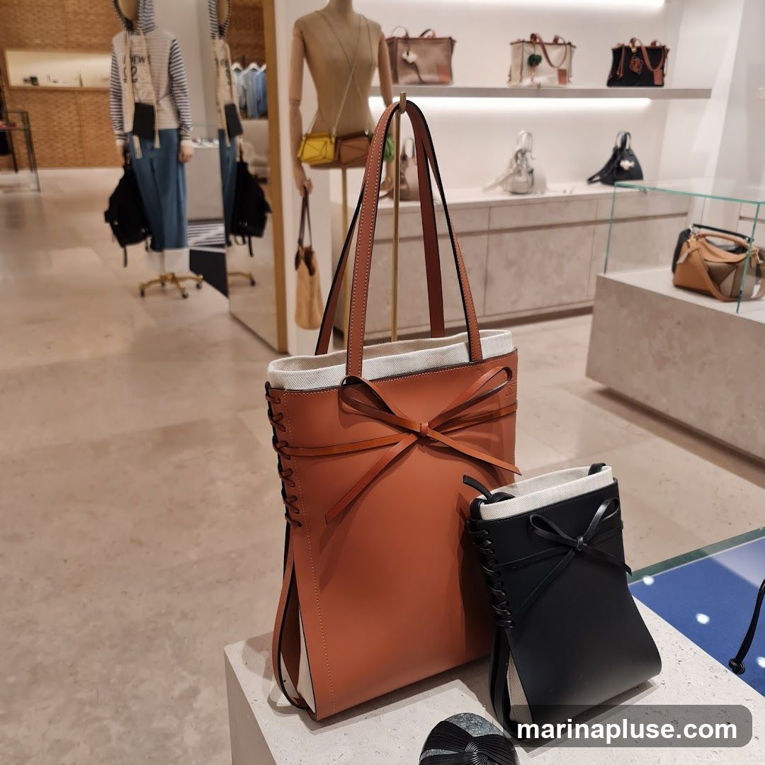 A display of various handbags in a retail store. In the foreground, two handbags are prominently featured: a larger tan tote bag with a bow detail and a smaller black clutch with a similar bow. In the background, mannequins are dressed in clothing, and shelves display more handbags and accessories. The store appears to be well-lit and organized, showcasing luxury goods.