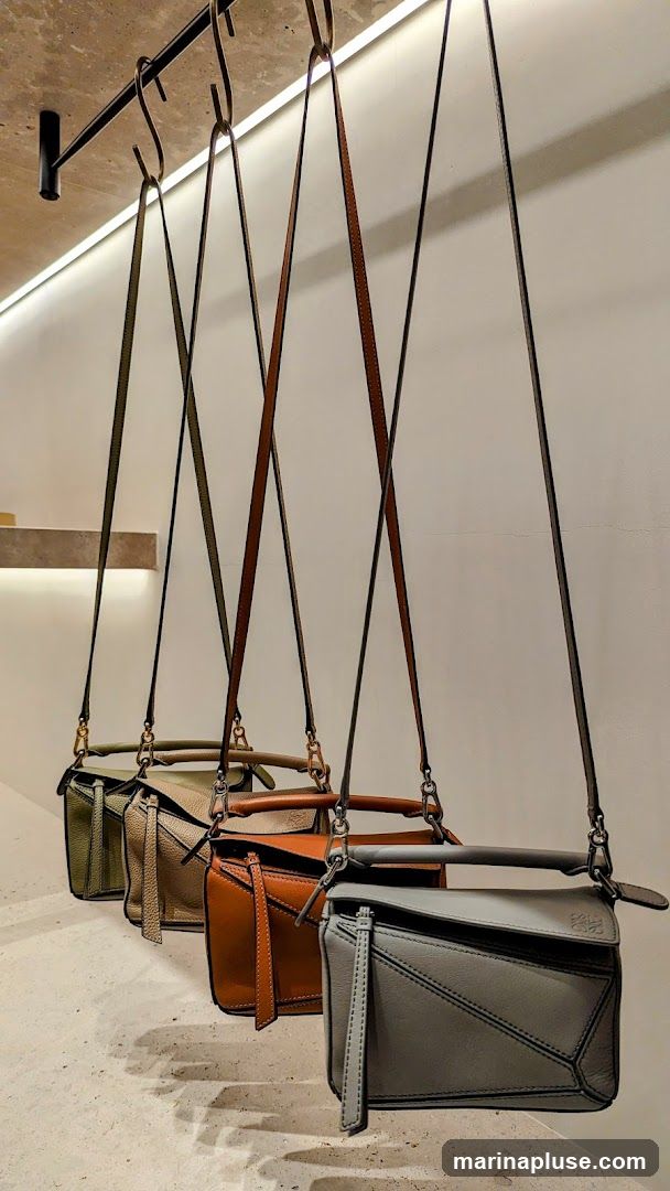 Four leather handbags in different colors (green, tan, orange, and grey) are displayed hanging from a rack. The grey bag in the foreground has a visible logo.