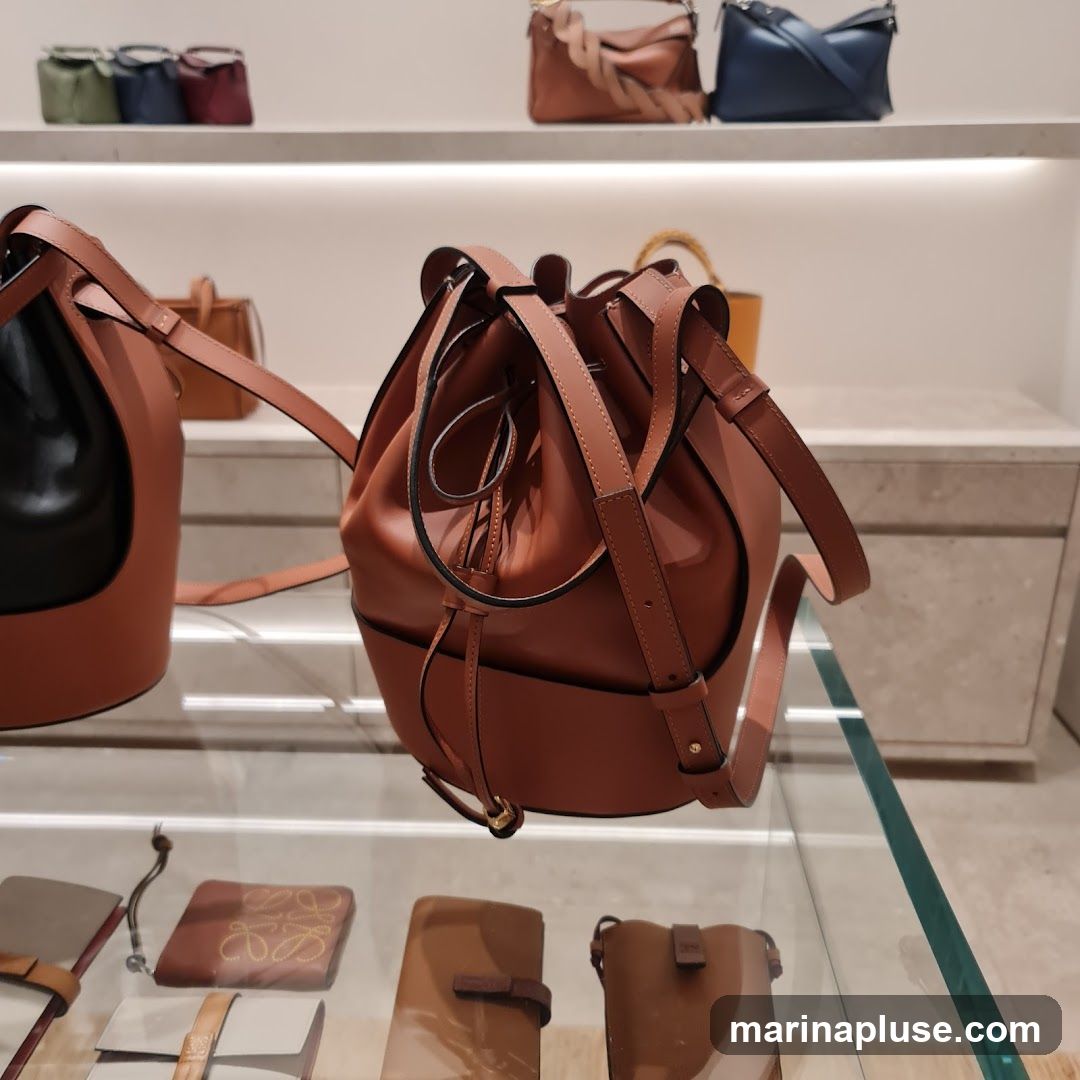 A close-up shot of several leather handbags displayed on shelves and in a glass case. The focus is on a large, brown drawstring bucket bag in the foreground. In the background, smaller handbags in various colors and styles are visible on shelves. The display appears to be in a retail store setting.