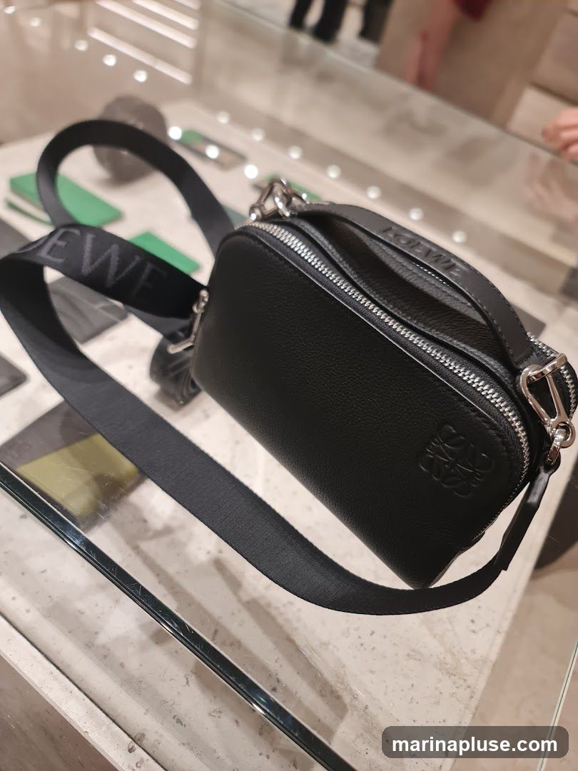 A black pebbled leather crossbody bag with a black strap featuring the LOEWE logo in white. The bag has silver-tone hardware and zippers. It is displayed on a glass surface in a retail setting.