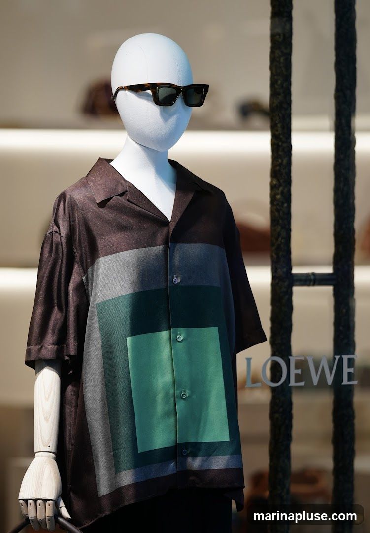A mannequin in a store window display wears a short-sleeved collared shirt with a geometric color-block pattern in shades of brown, gray, and green. The mannequin is also wearing dark sunglasses. The Loewe logo is visible on the glass window.