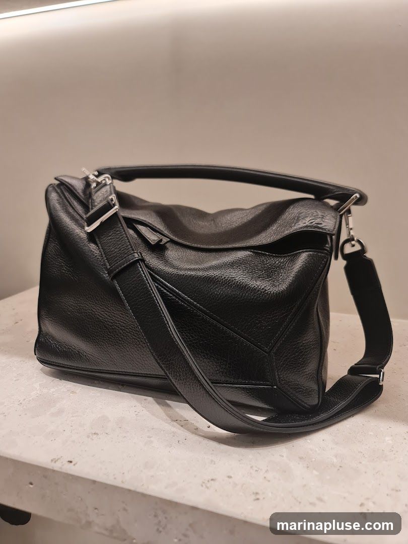 A black pebbled leather handbag with a top handle and a detachable shoulder strap is displayed on a textured surface. The bag has a geometric design on the front and silver-toned hardware.