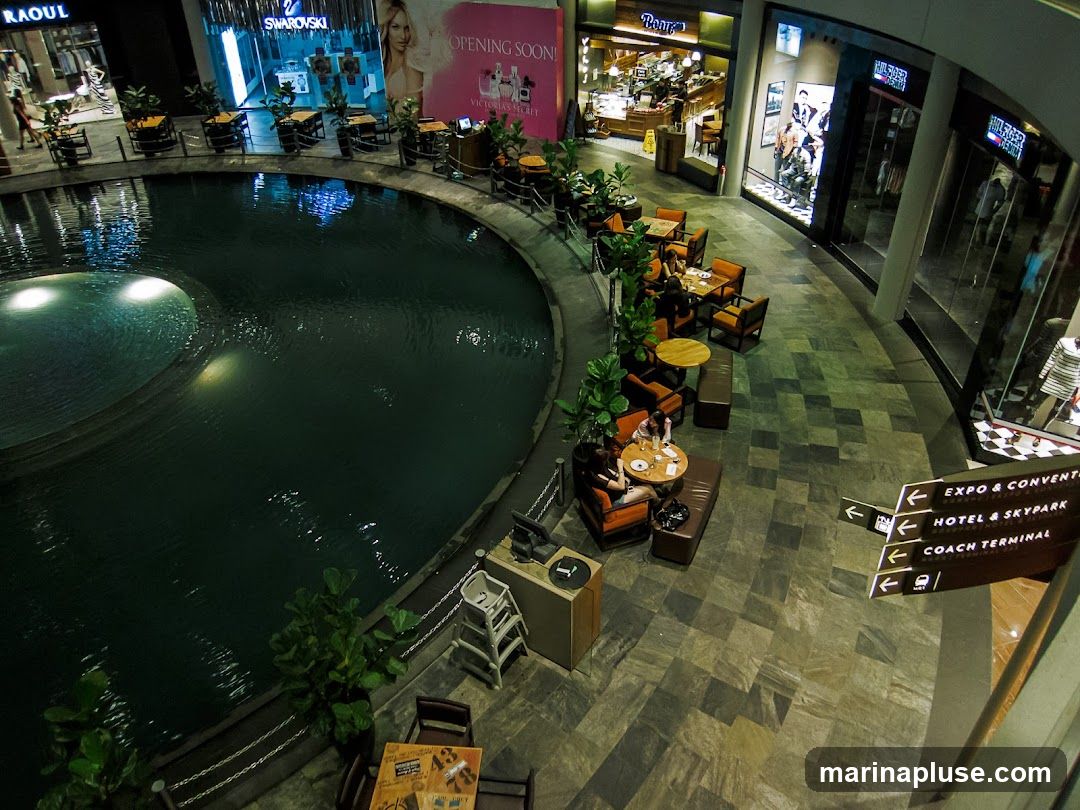 An aerial view of an indoor shopping mall featuring a large, circular water feature. Surrounding the water feature are various shops and seating areas with tables and chairs. Signage indicates directions to