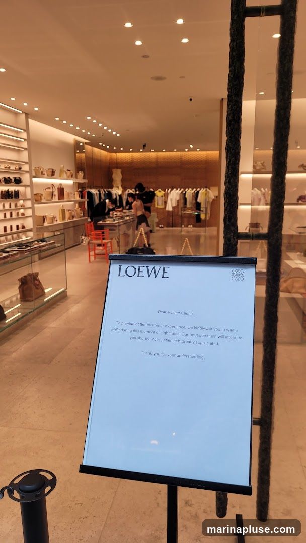 A sign inside a Loewe boutique informs clients that they may need to wait due to high traffic. The sign is white with black text and the Loewe logo. The background shows the interior of the store with clothing racks and displays.