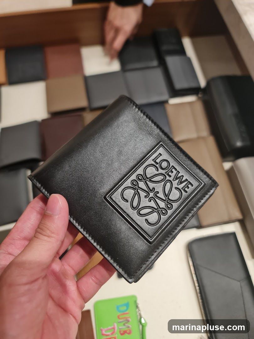 A hand holds a black leather bifold wallet with an embossed Loewe logo. In the background, various other wallets and small leather goods are displayed in wooden compartments. A small green pouch with the word "DUMB" is visible at the bottom.