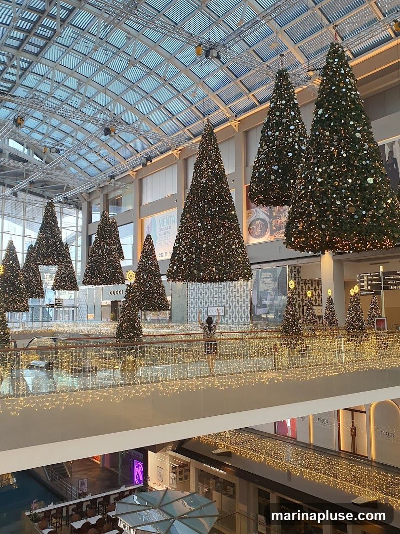 A wide shot of a shopping mall interior decorated for Christmas. Several large, decorated Christmas trees are suspended from the ceiling, while smaller trees line the railings of the upper levels. The mall features a glass roof and is adorned with numerous fairy lights, creating a festive atmosphere. A person is visible on an upper level walkway, facing away from the camera. Below, shops and dining areas are visible.