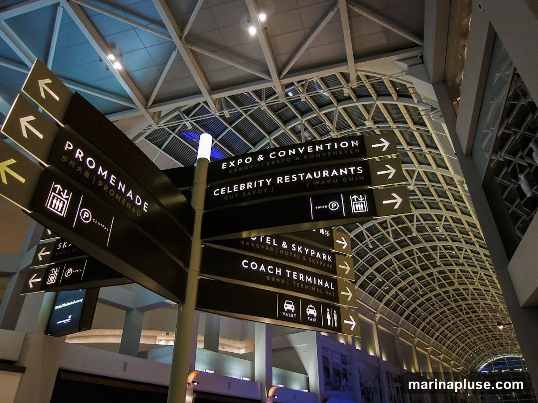 A multi-directional signpost with illuminated text and arrows points towards various locations within a large, modern building with a distinctive arched ceiling structure. Signs indicate 'PROMENADE', 'EXPO & CONVENTION', 'CELEBRITY RESTAURANTS', 'HOTEL & SKYPARK', and 'COACH TERMINAL', with some text in Chinese and other languages. Icons for parking, elevators, valet, and taxi services are also visible.