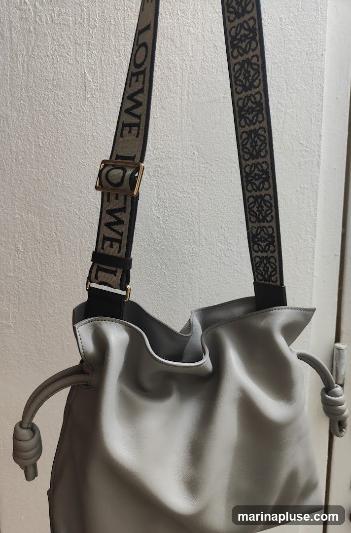 A light grey leather Loewe bag with a drawstring closure and knotted details on the sides. The bag has a wide strap with repeating Loewe logos and the word "LOEWE" printed on it.