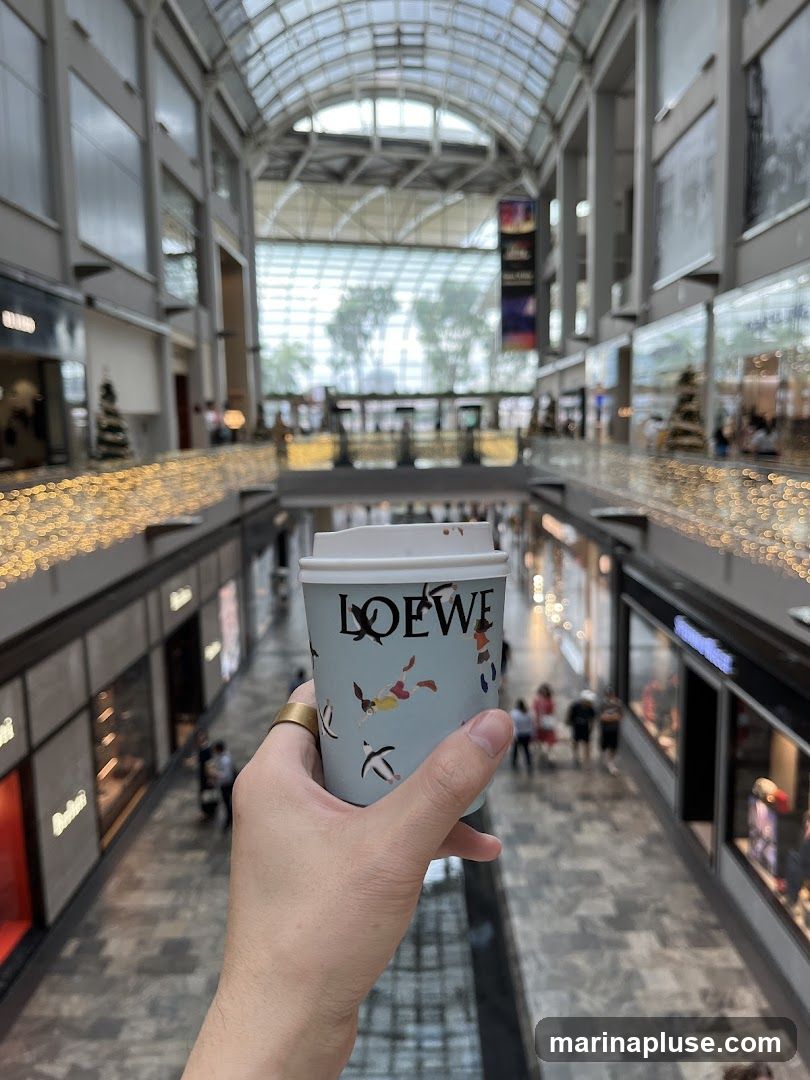 A hand holds a light blue paper coffee cup with the LOEWE logo and a whimsical pattern of figures and animals. The cup is in the foreground, with a blurred view of a modern, multi-level shopping mall in the background. The mall features a glass ceiling, escalators, and storefronts decorated with festive lights. People are visible walking on the lower levels.