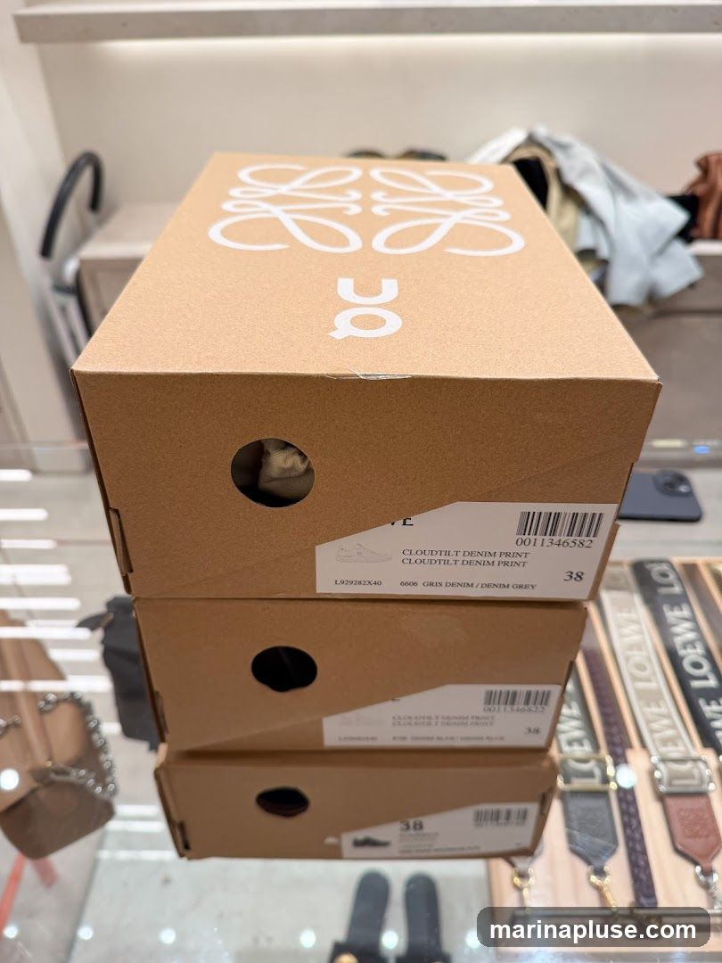 Three cardboard shoe boxes are stacked on top of each other. The top box has the Loewe logo and the 'On' logo printed in white. Labels on the boxes indicate shoe details such as 'CLOUDTILT DENIM PRINT' and size '38'. In the background, shelves with clothing and accessories are visible, including straps with the Loewe logo.