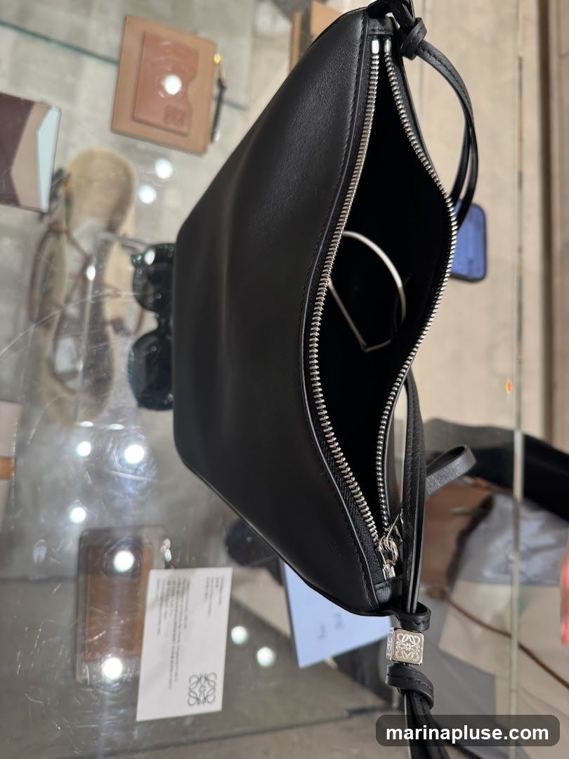 A black leather pouch with a silver zipper is displayed on a glass surface. The pouch is open, revealing a silver object inside. A small silver charm with a logo is attached to the zipper pull. In the background, other items are visible on display, including what appears to be a wallet and sunglasses. A tag with a logo is also visible on the glass surface.