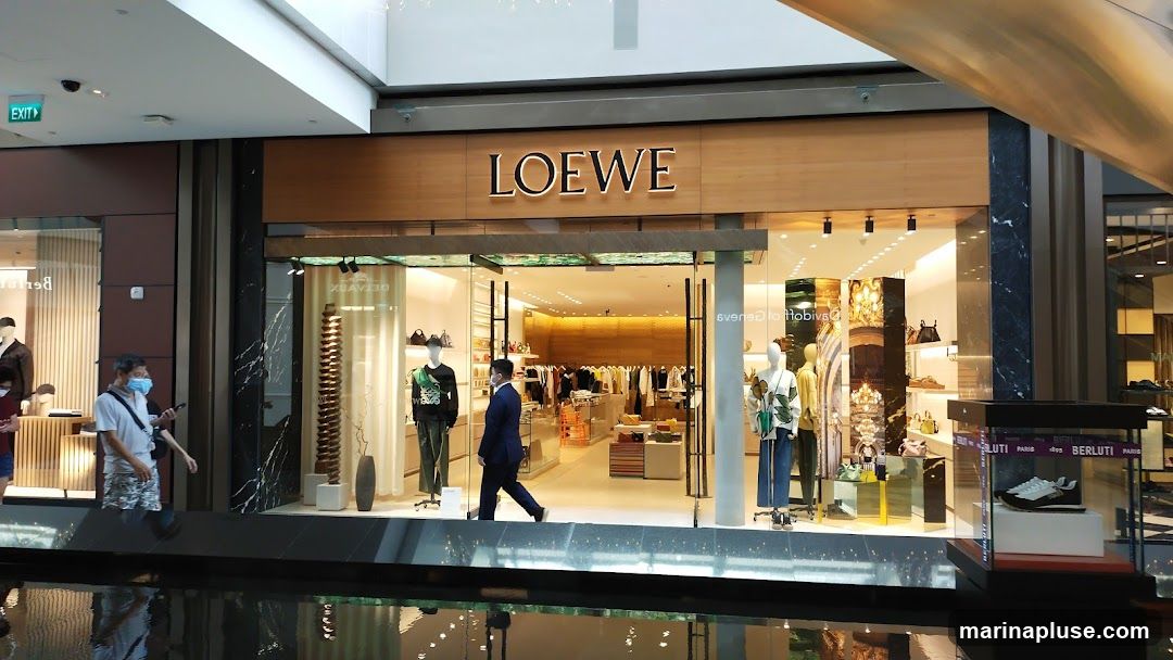 The exterior of a Loewe store with its name prominently displayed above the entrance. The store features large glass windows showcasing mannequins dressed in clothing and accessories, as well as displays of handbags and shoes. Inside, racks of clothing and shelves filled with merchandise are visible. Two individuals are walking past the storefront.