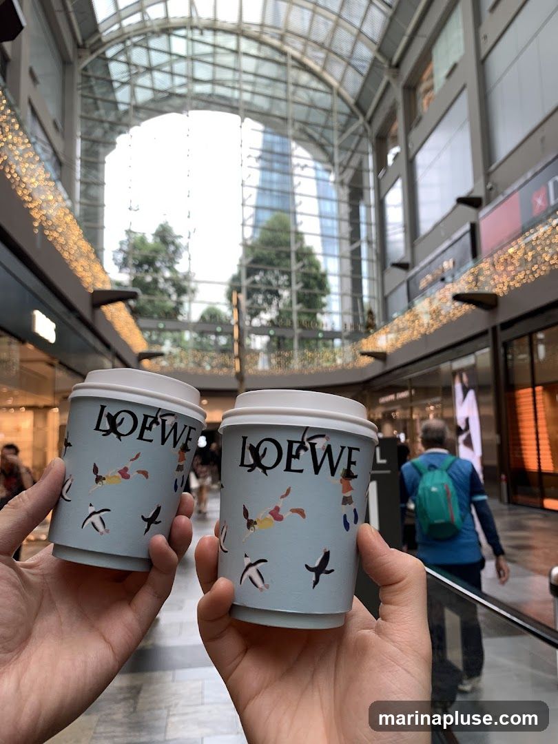 Two light blue disposable coffee cups with white lids are held up in the foreground. Both cups feature the black Loewe logo and a repeating pattern of cartoon figures and birds. The background shows a blurred interior of a modern shopping mall with a glass ceiling, escalators, and other shoppers.