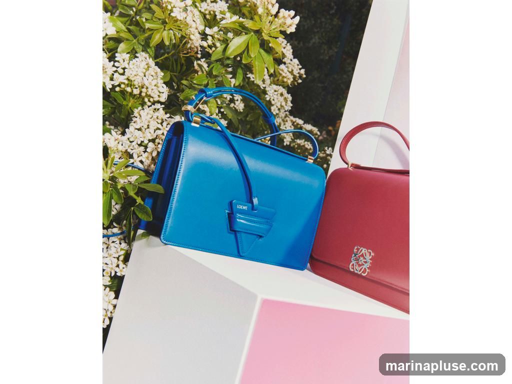 A bright blue handbag with a leather charm and a red handbag with a silver emblem are displayed on white and pink geometric pedestals. White flowers and green foliage are visible in the background.
