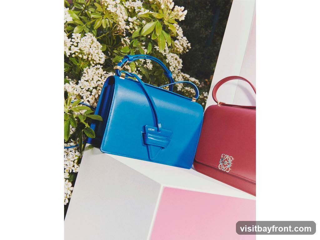 A bright blue handbag with a leather charm and a red handbag with a silver emblem are displayed on white and pink geometric pedestals. White flowers and green foliage are visible in the background.