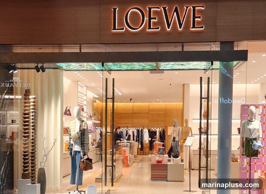 The exterior of a Loewe store is visible, with the brand name illuminated above the entrance. The storefront features large glass doors and windows, showcasing mannequins dressed in clothing and displaying handbags. Inside, racks of clothing and more handbags are arranged on shelves and displays. The interior has a warm, modern aesthetic with textured walls and bright lighting.