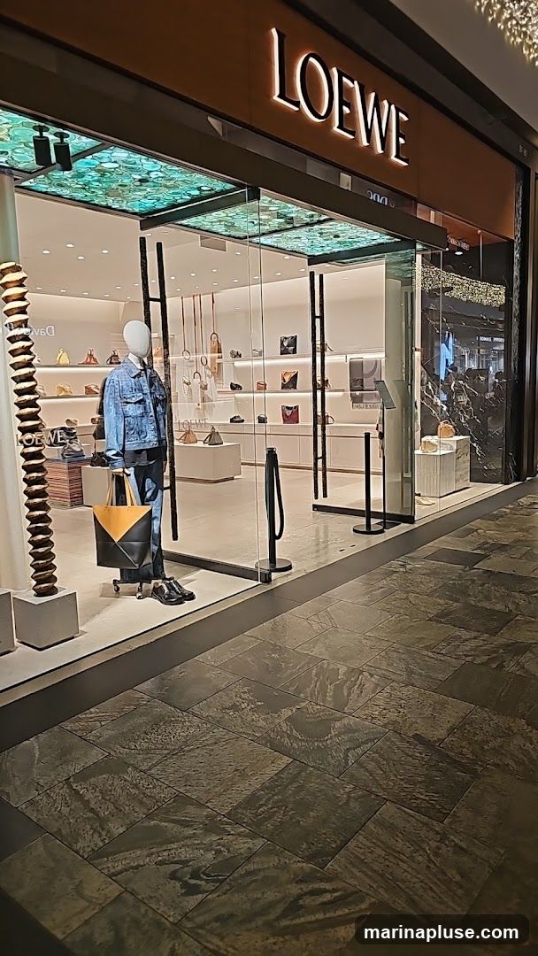 The exterior of a Loewe store with a large glass facade. A mannequin dressed in a denim jacket and jeans stands in the window display, holding a large tote bag. Shelves inside display various handbags and accessories. The Loewe logo is prominently displayed above the entrance.