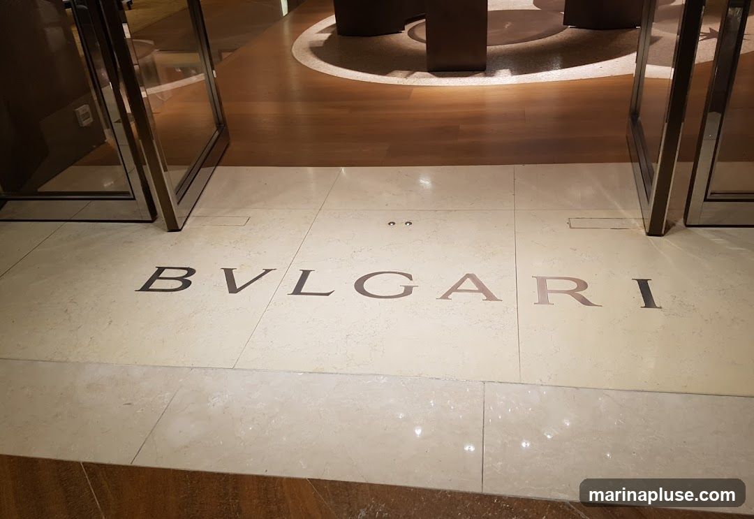 The image shows the BVLGARI logo inlaid into a marble floor, likely at the entrance of a store. The logo is spelled out in large, metallic letters. The floor transitions to a polished wooden surface further into the space, with circular patterns visible. The entrance appears to be framed by modern, metallic door frames and glass panels.
