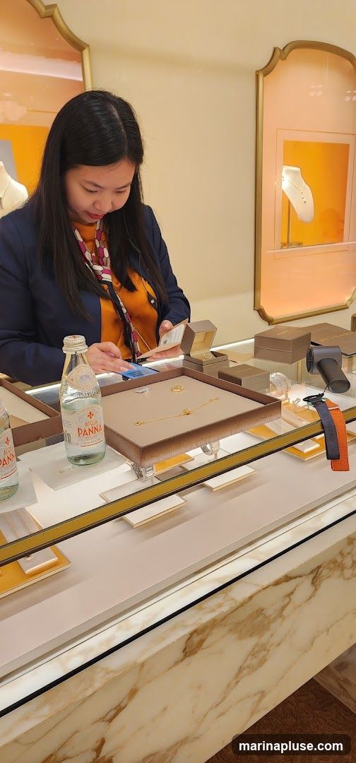A woman in a dark blazer and an orange patterned blouse is looking at a small card and her phone while standing behind a jewelry display case. The display case contains several pieces of gold jewelry, including necklaces and bracelets, as well as jewelry boxes. Two bottles of Acqua Panna water are also visible on the counter. In the background, a display case shows a necklace on a mannequin. The store has a luxurious feel with marble accents and soft lighting.