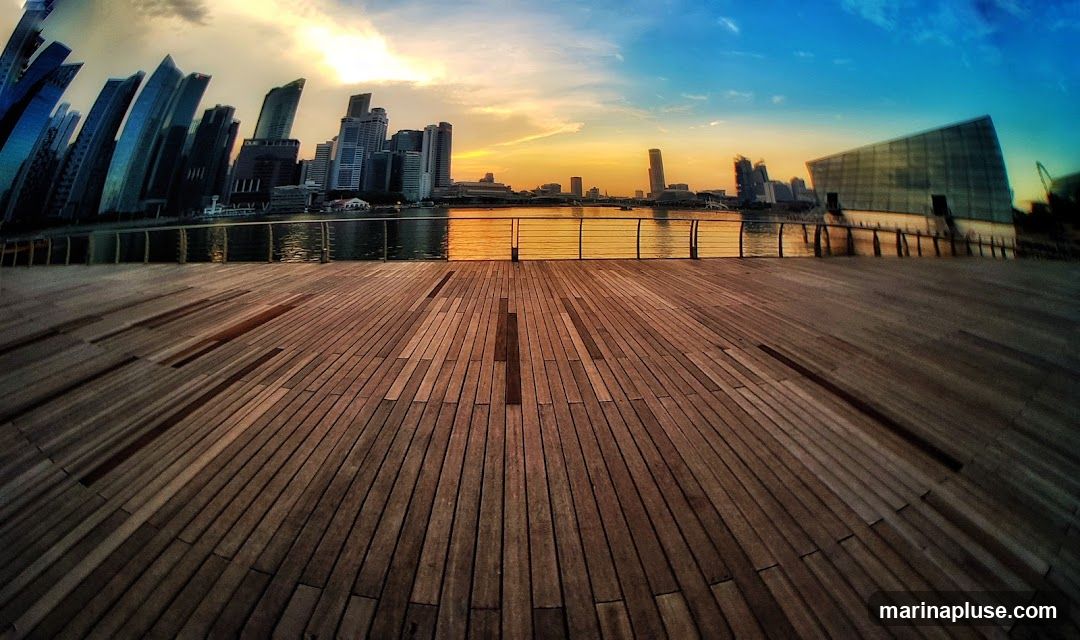 A wide-angle view of a modern cityscape at sunset, with a wooden boardwalk in the foreground. The sky transitions from warm orange and yellow hues near the horizon to a deep blue overhead. Silhouetted skyscrapers rise in the distance, reflected in the calm water. A contemporary building with a glass facade is visible on the right. The wooden planks of the boardwalk are laid out in a pattern, with shadows creating a sense of depth.