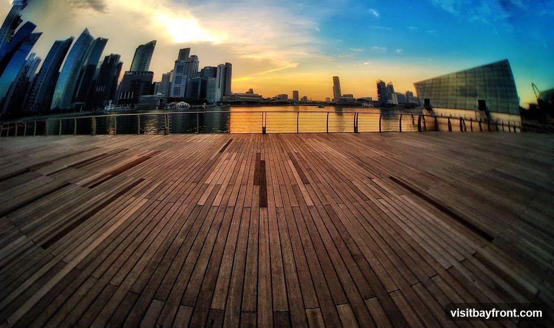 A wide-angle view of a modern cityscape at sunset, with a wooden boardwalk in the foreground. The sky transitions from warm orange and yellow hues near the horizon to a deep blue overhead. Silhouetted skyscrapers rise in the distance, reflected in the calm water. A contemporary building with a glass facade is visible on the right. The wooden planks of the boardwalk are laid out in a pattern, with shadows creating a sense of depth.