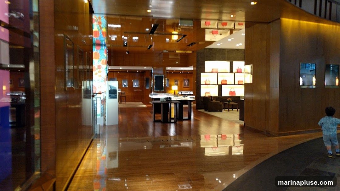 A wide shot of the interior of a luxury retail store. The floor is polished wood, reflecting the warm lighting and displays. In the foreground, a young child walks away from the camera. The store features display cases with handbags and other accessories illuminated by soft lighting. The overall ambiance is elegant and spacious.
