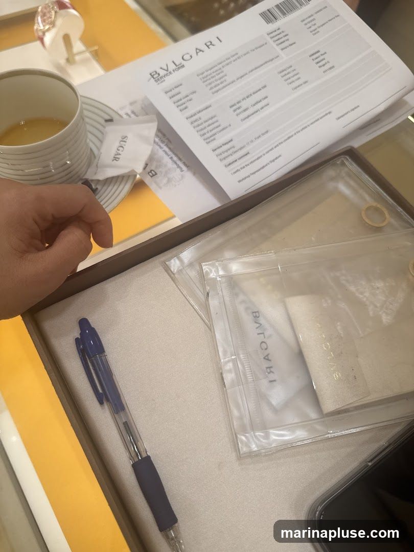 A BVLGARI service form is laid out on a counter, next to a cup of tea and a sugar packet. A blue pen rests in a box containing several clear plastic pouches with BVLGARI branding. A watch is visible in the background.