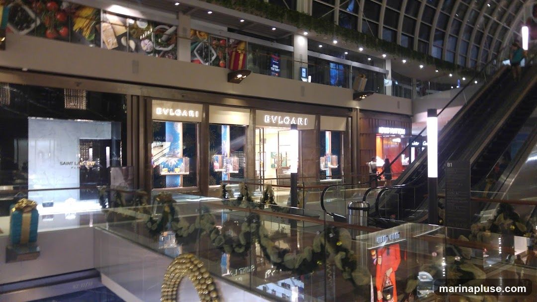The exterior of a Bulgari store is visible, with its name illuminated above the entrance. The store features large display windows showcasing jewelry and other luxury items. To the left, a Saint Laurent store is also visible. The scene is set within a shopping mall, with escalators and other shops in the background.