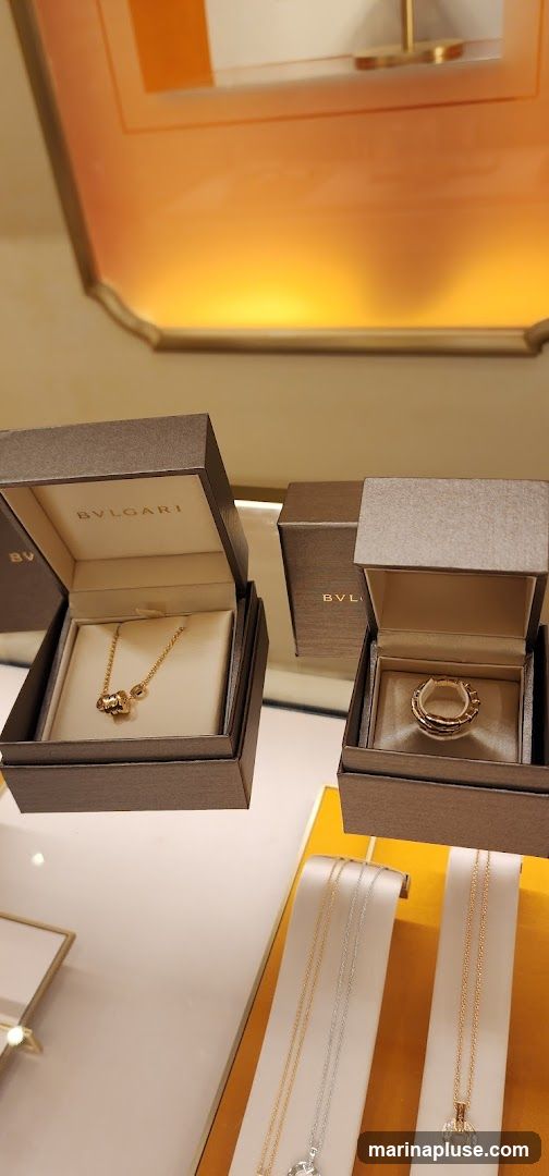 A display of luxury jewelry, featuring necklaces and rings in open boxes and on stands. The brand name BVLGARI is visible on the interior of the boxes. The lighting is warm and highlights the gold and diamond pieces.
