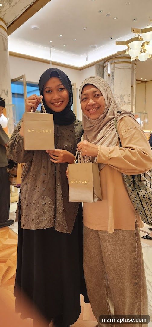Two women, both wearing hijabs, are smiling and holding shopping bags with the Bulgari logo. The woman on the left is wearing a dark grey embroidered top and a black skirt. The woman on the right is wearing a light brown top and brown pants. They are inside a store with marble columns and a chandelier visible in the background.