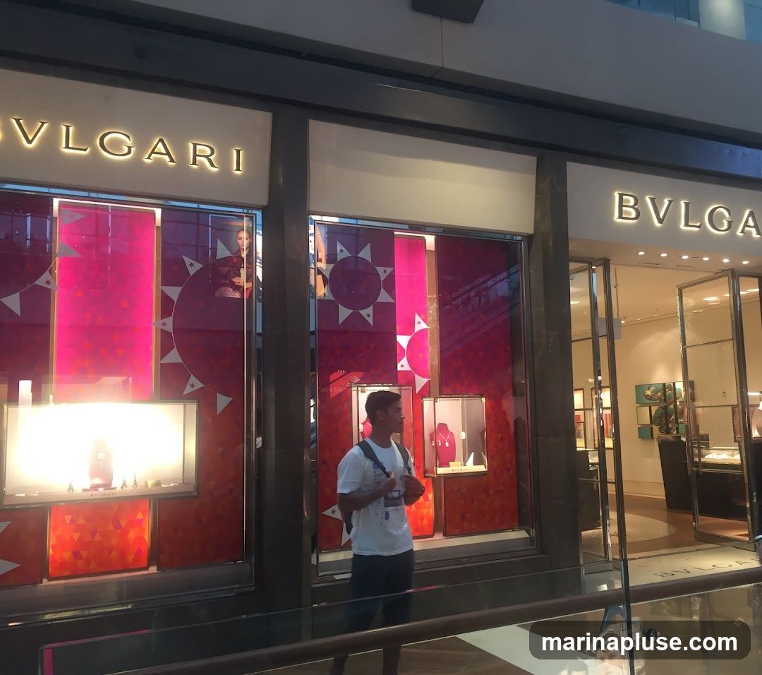 The exterior of a Bulgari store with illuminated signage above the entrance. The storefront features large display windows showcasing jewelry and mannequins. A young man stands in front of the window display.