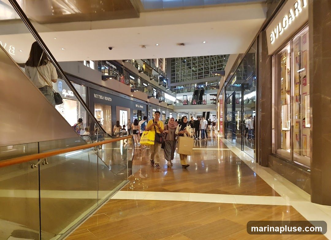 A wide shot of a modern shopping mall interior with polished wooden floors and bright lighting. People are walking along the corridor, some carrying shopping bags. Storefronts of luxury brands like Bottega Veneta and Bvlgari are visible on either side. Escalators and multiple levels of shops can be seen in the background.