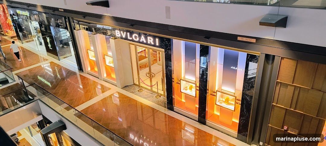 The exterior of a Bulgari jewelry store in a mall. The storefront features the Bulgari logo illuminated above the entrance, with large display windows showcasing jewelry. The entrance has ornate gold doors and velvet ropes. The surrounding mall interior is visible, with polished floors and other store fronts.