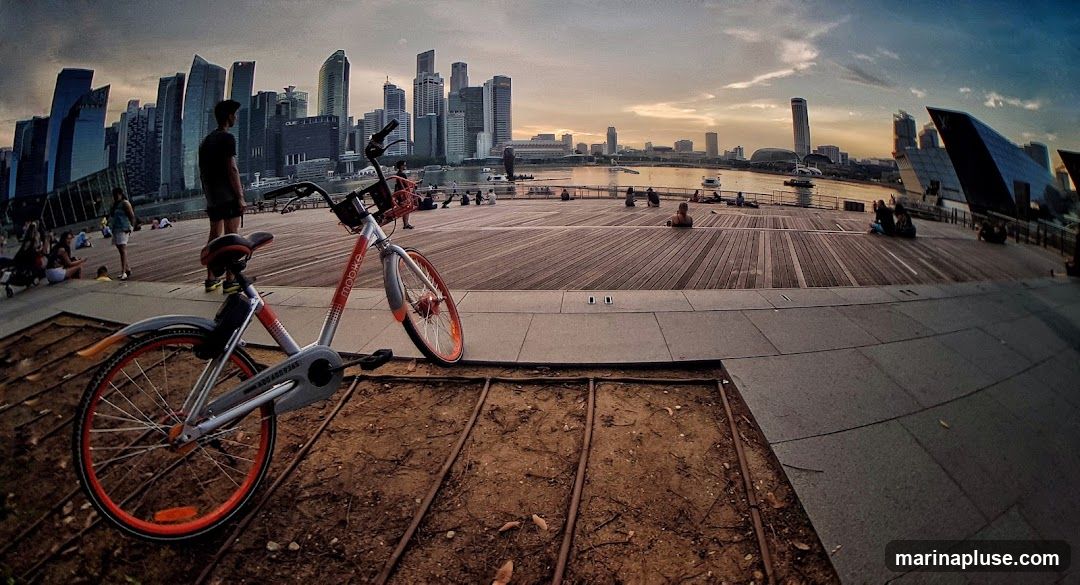 A shared bicycle with orange accents is parked on a wooden promenade. In the background, a modern city skyline with numerous skyscrapers is visible across a body of water. Several people are seated and relaxing on the promenade, enjoying the view during what appears to be sunset or dusk. The sky is a mix of blue and orange hues.