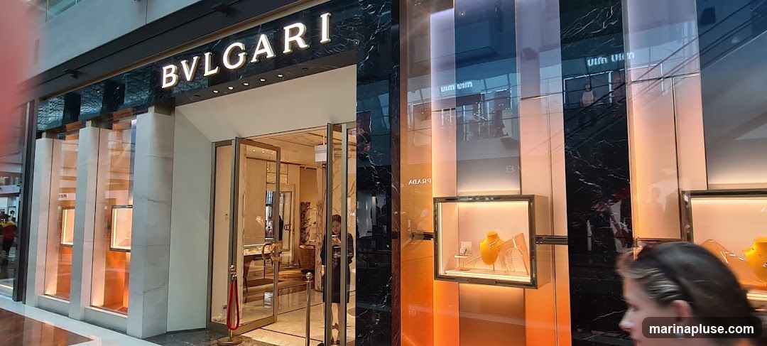 The exterior of a BVLGARI store with a prominent sign above the entrance. The storefront features large glass doors and display windows showcasing jewelry. The facade is decorated with marble and illuminated accents.