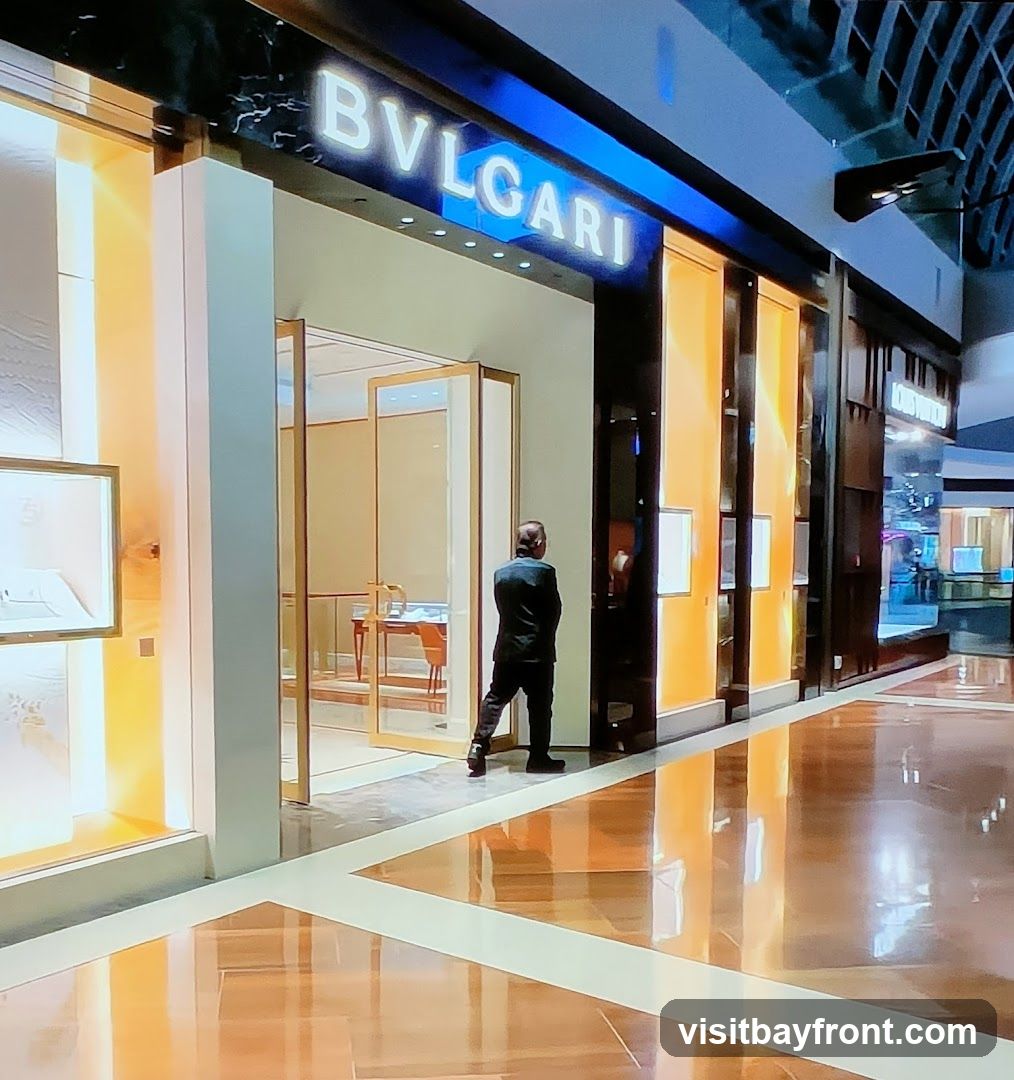 A man stands at the entrance of a Bvlgari store. The store has a modern facade with illuminated display windows and the Bvlgari logo prominently displayed above the entrance. The interior is visible through the glass doors, showing display cases and furniture. The floor is highly reflective, mirroring the store's lights and facade.