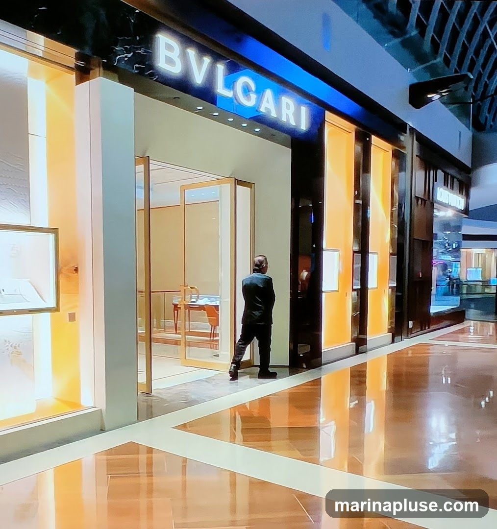 A man stands at the entrance of a Bvlgari store. The store has a modern facade with illuminated display windows and the Bvlgari logo prominently displayed above the entrance. The interior is visible through the glass doors, showing display cases and furniture. The floor is highly reflective, mirroring the store's lights and facade.