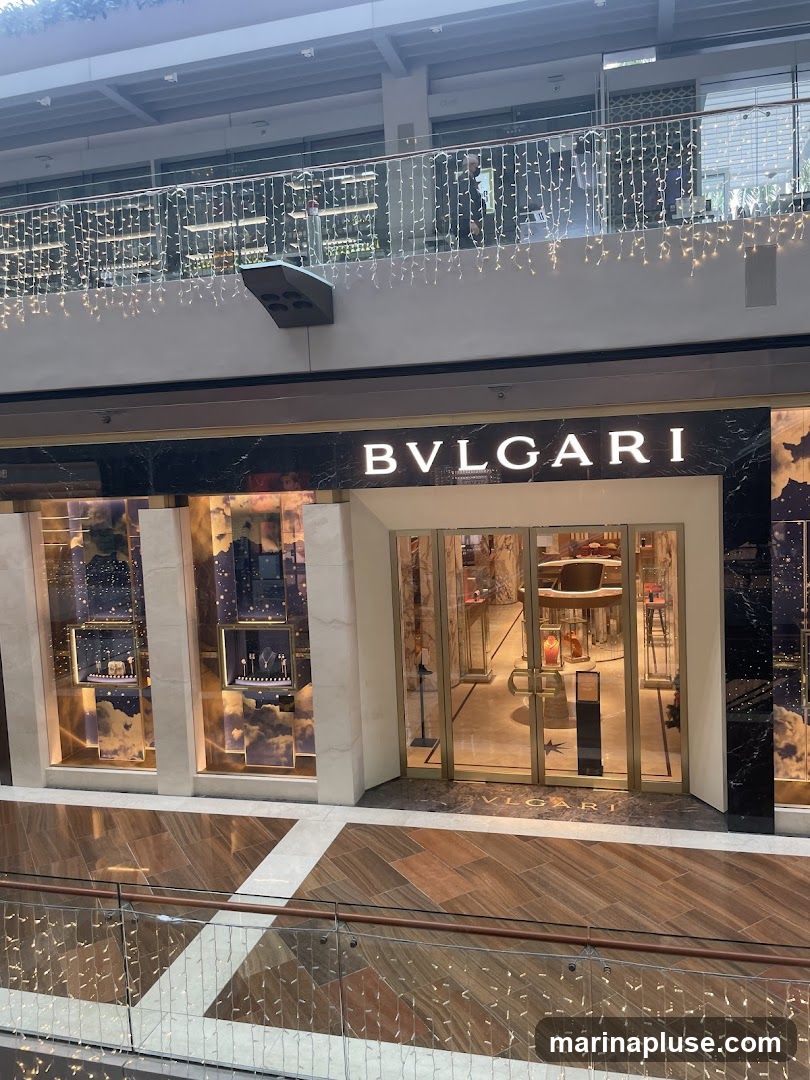 The exterior of a BVLGARI store with its name prominently displayed above the entrance. The store features large glass windows showcasing jewelry displays and a polished interior. The facade is made of dark marble and accented with gold trim.