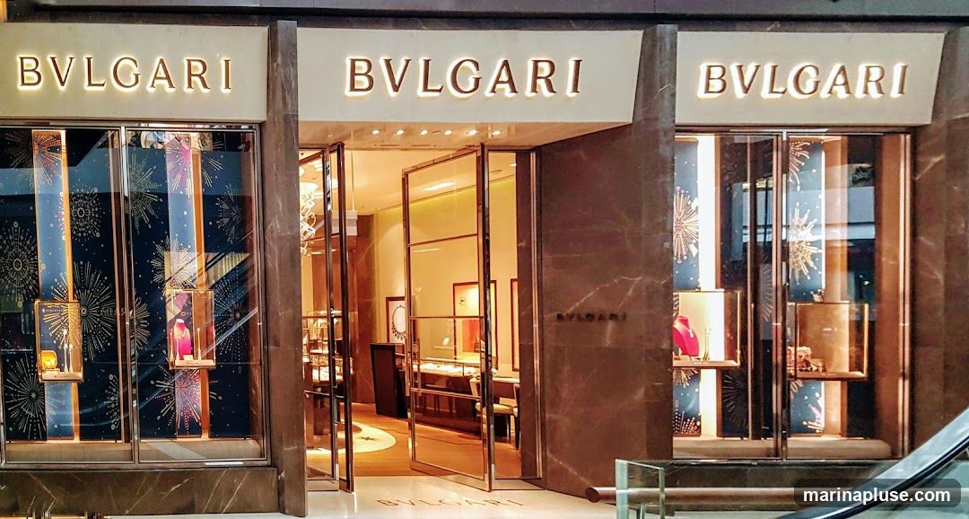 The exterior of a Bulgari store with illuminated signage above the entrance. The storefront features large glass windows displaying jewelry and decorative elements. The entrance has double glass doors, and the building facade is made of dark marble.