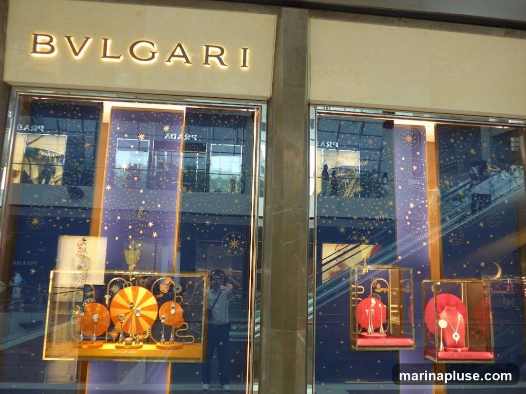 The exterior of a Bulgari store with illuminated signage above the entrance. The display windows feature a dark blue background with gold stars and crescent moons. Inside the windows, jewelry is displayed on red velvet stands. One display case shows several orange circular objects with smaller items in the center, and another shows necklaces and earrings. A person is visible taking a photo of their reflection in the window.
