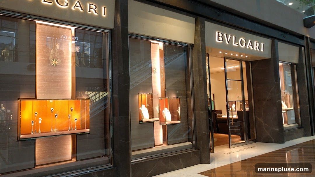 The exterior of a Bulgari store with illuminated signage above the entrance. The storefront features large display windows showcasing jewelry and watches. The entrance is open, revealing the interior of the store with display cases and lighting.