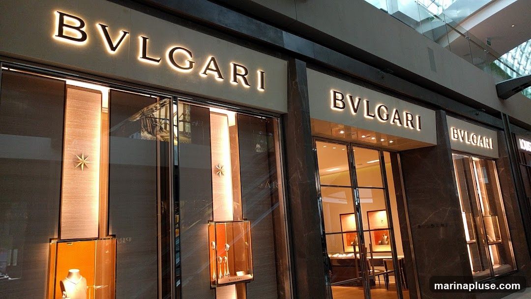 The exterior of a Bvlgari store with illuminated signage above the entrance. The storefront features large glass windows displaying jewelry in illuminated cases. The facade is made of dark marble and brushed metal accents.