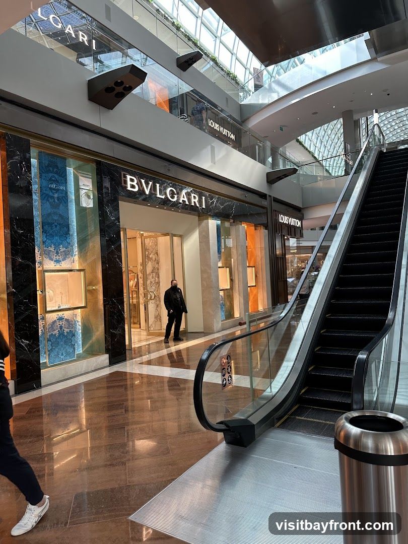 An interior view of a modern shopping mall featuring luxury brand stores. The prominent Bulgari store is visible with its distinctive signage and marble facade. Adjacent to it, Louis Vuitton stores are also present on multiple levels. An escalator runs alongside the stores, and the mall's architecture includes a glass ceiling allowing natural light to filter in. A person stands near the entrance of the Bulgari store, and another person walks past the escalator.