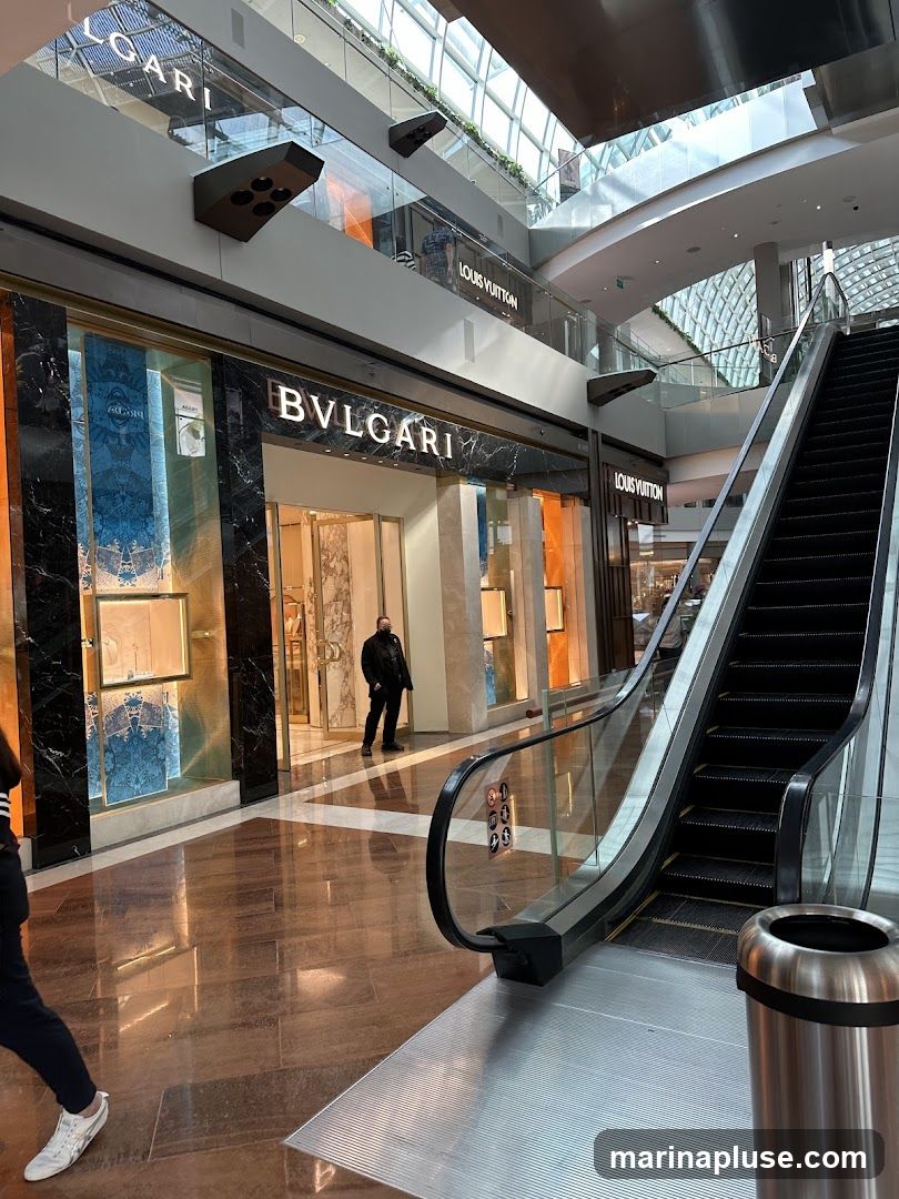An interior view of a modern shopping mall featuring luxury brand stores. The prominent Bulgari store is visible with its distinctive signage and marble facade. Adjacent to it, Louis Vuitton stores are also present on multiple levels. An escalator runs alongside the stores, and the mall's architecture includes a glass ceiling allowing natural light to filter in. A person stands near the entrance of the Bulgari store, and another person walks past the escalator.
