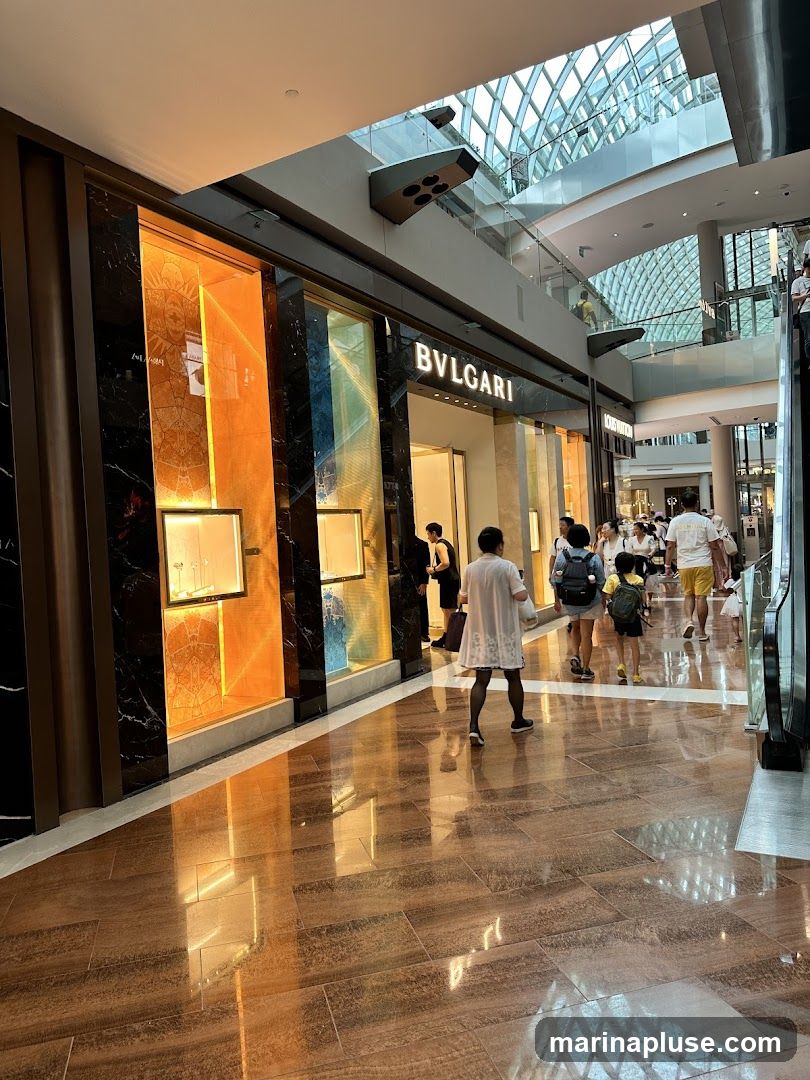 The exterior of a Bvlgari store in a modern shopping mall. The storefront features a dark marble facade with illuminated display windows showcasing jewelry. The Bvlgari logo is prominently displayed above the entrance. People are walking by the store and on escalators in the background.