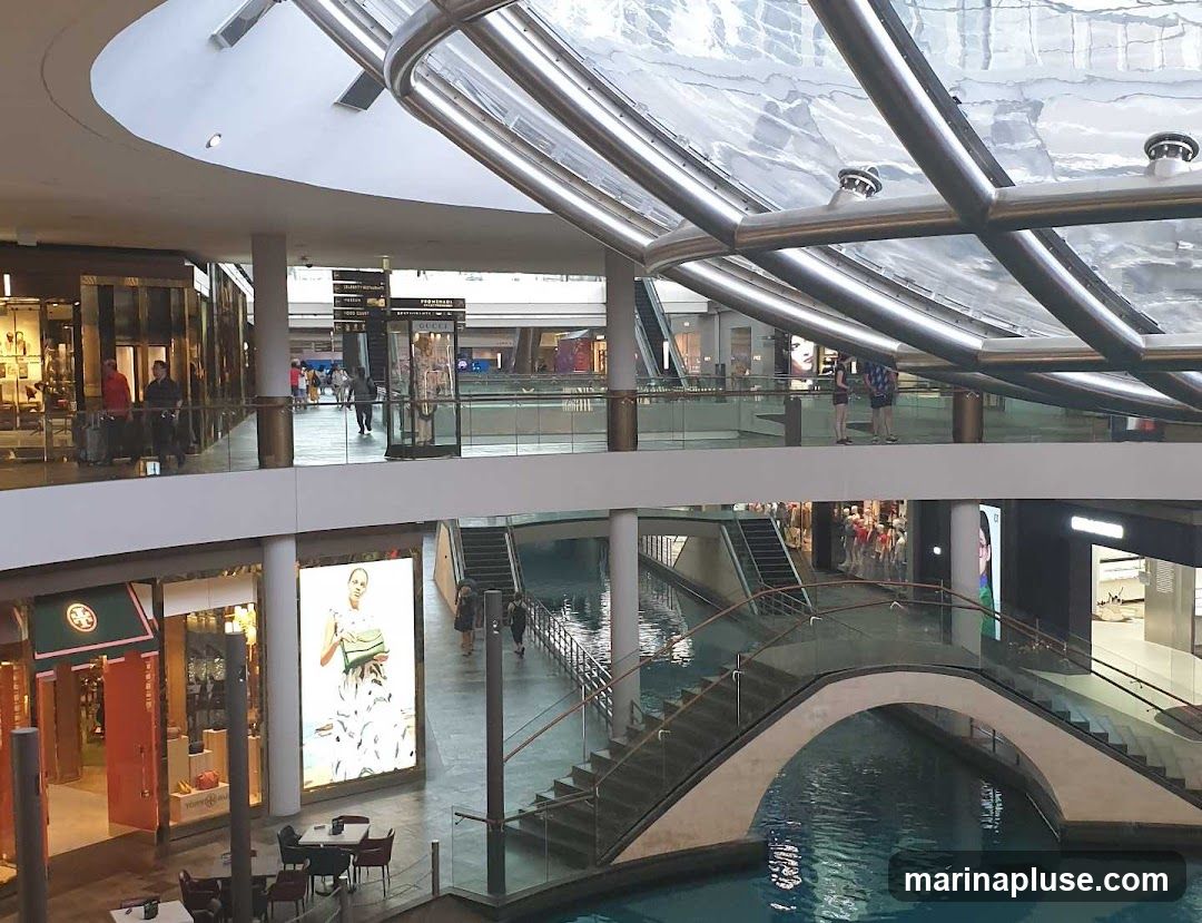 The image shows the interior of a multi-level shopping mall. A canal runs through the lower level, crossed by a decorative bridge. Escalators and walkways connect the different floors. Various shops are visible, including one with a large digital display featuring a model. People are seen walking around the mall. The architecture features a modern design with a glass and metal roof structure.
