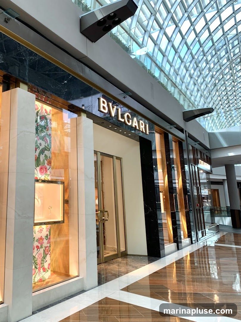 The exterior of a Bulgari store with its name prominently displayed above the entrance. The storefront features large display windows showcasing jewelry and decorative floral wallpaper. The entrance has glass doors with gold-colored handles. The building is located in a modern indoor shopping mall with a distinctive glass ceiling.