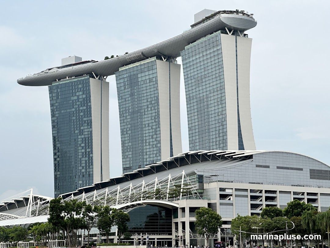 A wide shot of the Marina Bay Sands hotel in Singapore. The iconic building features three towers connected by a large rooftop deck. The towers are clad in glass and white panels. In the foreground, there is a large, modern convention center with a curved glass roof. Trees and a few people are visible at the base of the buildings. The sky is overcast.